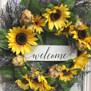 Pu&ograve; includere: Una ghirlanda di girasoli con un cartello di benvenuto. La ghirlanda &egrave; fatta di girasoli, carciofi e verde. Il cartello &egrave; bianco con lettere nere.
