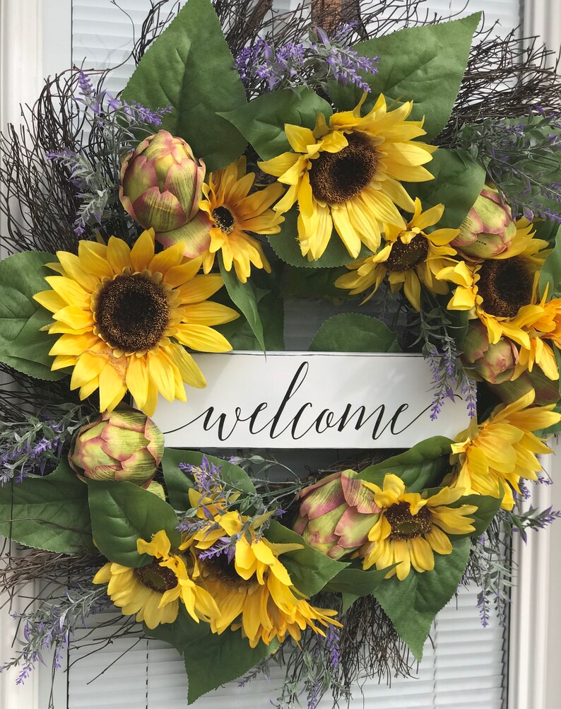 Pu&ograve; includere: Una ghirlanda di girasoli con un cartello di benvenuto. La ghirlanda &egrave; fatta di girasoli, carciofi e verde. Il cartello &egrave; bianco con lettere nere.