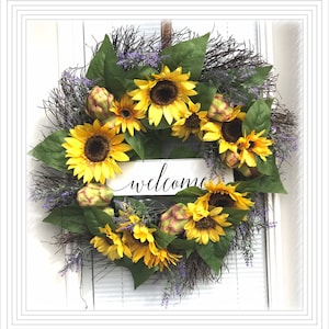 Pu&ograve; includere: Una ghirlanda di girasoli con un cartello di benvenuto. La ghirlanda &egrave; fatta di girasoli artificiali, carciofi e verde. Il cartello &egrave; bianco con lettere nere.