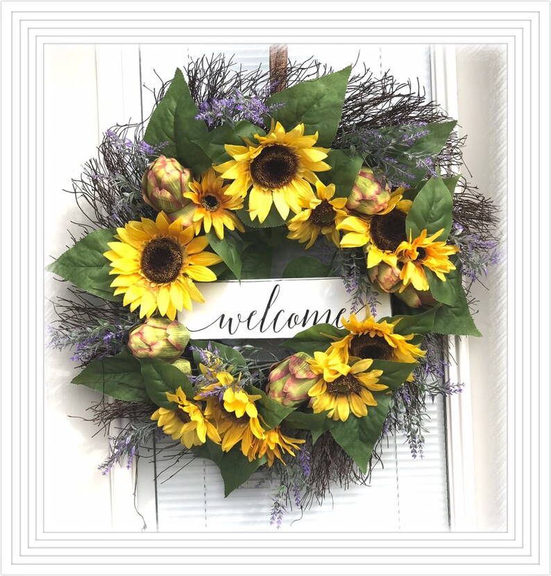 Pu&ograve; includere: Una ghirlanda di girasoli con un cartello di benvenuto. La ghirlanda &egrave; fatta di girasoli artificiali, carciofi e verde. Il cartello &egrave; bianco con lettere nere.