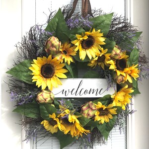 Pu&ograve; includere: Una ghirlanda di girasoli con un cartello di benvenuto. La ghirlanda &egrave; fatta di ramoscelli marroni, foglie verdi, fiori viola e girasoli gialli. Il cartello &egrave; bianco con lettere nere.