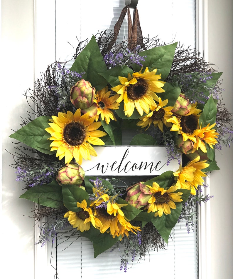 Pu&ograve; includere: Una ghirlanda di girasoli con un cartello di benvenuto. La ghirlanda &egrave; fatta di ramoscelli marroni, foglie verdi, fiori viola e girasoli gialli. Il cartello &egrave; bianco con lettere nere.