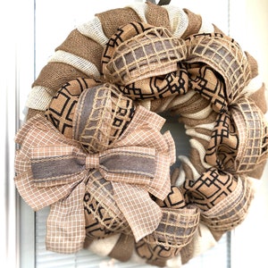 May include: A brown and beige burlap wreath with a large brown and beige plaid bow. The wreath is decorated with brown and beige ribbon and has a geometric pattern.