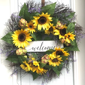 Pu&ograve; includere: Una ghirlanda di girasoli con un cartello di benvenuto. La ghirlanda &egrave; fatta di girasoli artificiali, verde e rametti di lavanda. Il cartello &egrave; bianco con lettere nere.