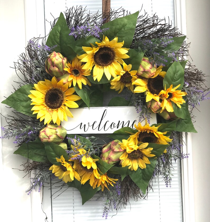 Pu&ograve; includere: Una ghirlanda di girasoli con un cartello di benvenuto. La ghirlanda &egrave; fatta di girasoli artificiali, verde e rametti di lavanda. Il cartello &egrave; bianco con lettere nere.