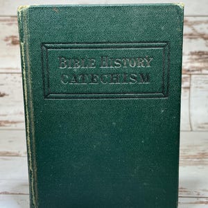 May include: A vintage green book titled "Bible History Catechism." The cover has a textured appearance with the title embossed in a rectangular frame. The book's edges show signs of wear, suggesting its age.