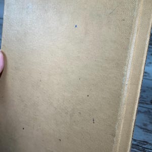 May include: A tan-colored book with a textured cover and a white spine. The book is slightly worn and has a few small dark spots on the cover.