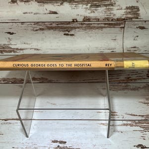 May include: A vintage children's book titled "Curious George Goes to the Hospital" by H.A. Rey. The book is yellow with black text and is displayed on a clear acrylic book stand.