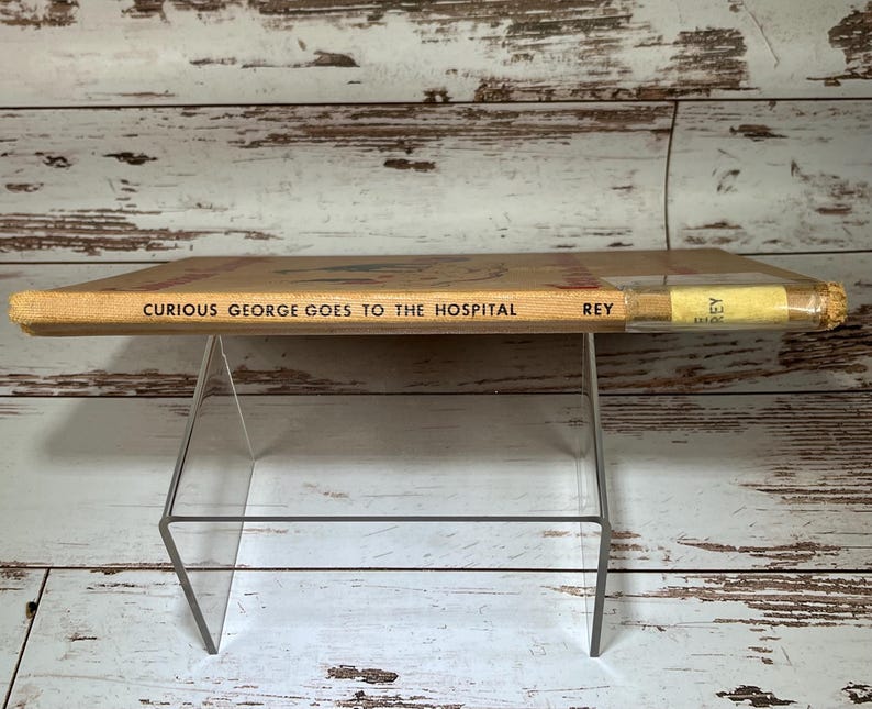 May include: A vintage children's book titled "Curious George Goes to the Hospital" by H.A. Rey. The book is yellow with black text and is displayed on a clear acrylic book stand.