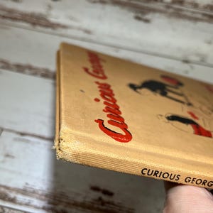 May include: A vintage hardcover book titled "Curious George" with a red and white illustration of Curious George on the cover. The book is in good condition with some wear and tear on the edges.