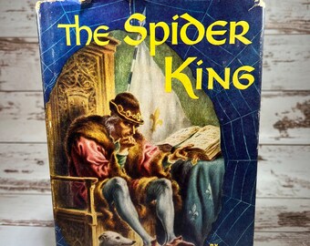 May include: A vintage hardcover book titled "The Spider King" by Lawrence Schoonover. The cover features a detailed illustration of a man in a crown and fur-trimmed robe sitting on a throne, reading a book. A small white dog sits at his feet. The background is a dark blue with a spiderweb pattern.