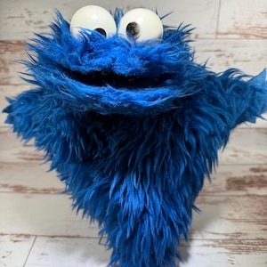 May include: A blue plush toy with long, shaggy faux fur, featuring two large, round, white plastic eyes with black pupils. The creature has a wide, open mouth and outstretched arms, covered entirely in vibrant blue synthetic fibers.