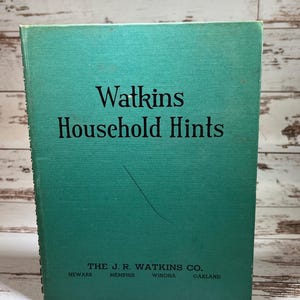 May include: A vintage teal book titled "Watkins Household Hints." The cover features black text and the company name, "The J.R. Watkins Co.," with city locations. The book's spine is visible, and the background is a light wood.