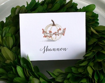 Könnte beinhalten: Ein weißer Teller mit einem grünen Buchsbaumkranz und einer weißen Karte mit einem handgemalten weißen Kürbis mit roten Beeren und dem Namen "Shannon" in schwarzer Kalligraphie.