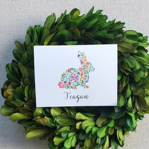 May include: A white card with the name "Teagan" in script, featuring a floral bunny design, rests on a green wreath. The bunny is composed of colorful flowers. The wreath is made of green leaves, set against a neutral background.