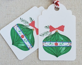 May include: Two white gift tags with a green and red Christmas ornament illustration. The tags are tied together with a red and white twine.