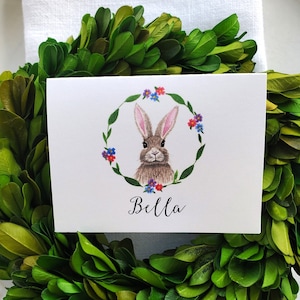 May include: A white card with a watercolor illustration of a brown bunny rabbit with pink ears and blue eyes. The bunny is surrounded by a wreath of green leaves and colorful flowers. The card has the name "Bella" written in cursive at the bottom.