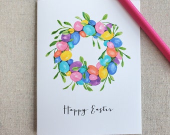 May include: A white card with a watercolor illustration of a wreath made of colorful Easter eggs and green leaves. The text "Happy Easter" is written in black cursive below the wreath.