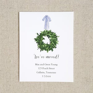 May include: A white card with a green wreath and a blue bow on top. The text "We've moved!" is written in cursive. Below the text is the address: Max and Gwen Young, 123 Peach Street, Gallatin, Tennessee 37066.