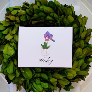 May include: A white card with a purple and white flower illustration in a cracked egg shell. The card has the name "Finley" written in black cursive.