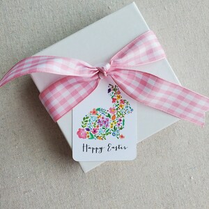 Puede incluir: Una caja de regalo blanca con una cinta de cuadros rosa y blanca atada en un lazo. Una etiqueta de regalo con un dise&ntilde;o de conejo floral y el texto "Feliz Pascua" est&aacute; unida a la caja.