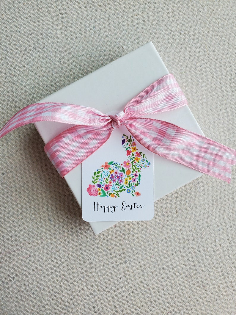 Puede incluir: Una caja de regalo blanca con una cinta de cuadros rosa y blanca atada en un lazo. Una etiqueta de regalo con un dise&ntilde;o de conejo floral y el texto "Feliz Pascua" est&aacute; unida a la caja.