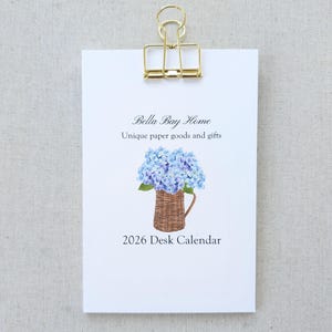May include: A white desk calendar clipped with a gold binder clip. The calendar features the text "Bella Bay Home" and "Unique paper goods and gifts." A watercolor illustration of blue hydrangeas in a woven basket is below the text. The calendar reads "2026 Desk Calendar."