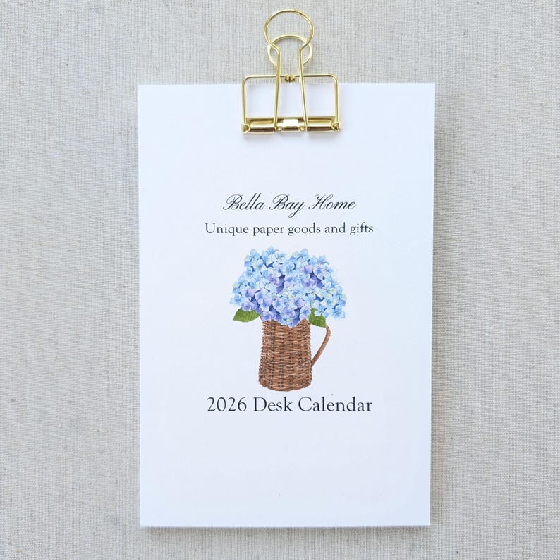 May include: A white desk calendar clipped with a gold binder clip. The calendar features the text "Bella Bay Home" and "Unique paper goods and gifts." A watercolor illustration of blue hydrangeas in a woven basket is below the text. The calendar reads "2026 Desk Calendar."