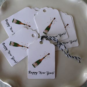 May include: A set of white gift tags with a black and white string. Each tag features a champagne bottle popping with the text "Happy New Year!" printed in black.