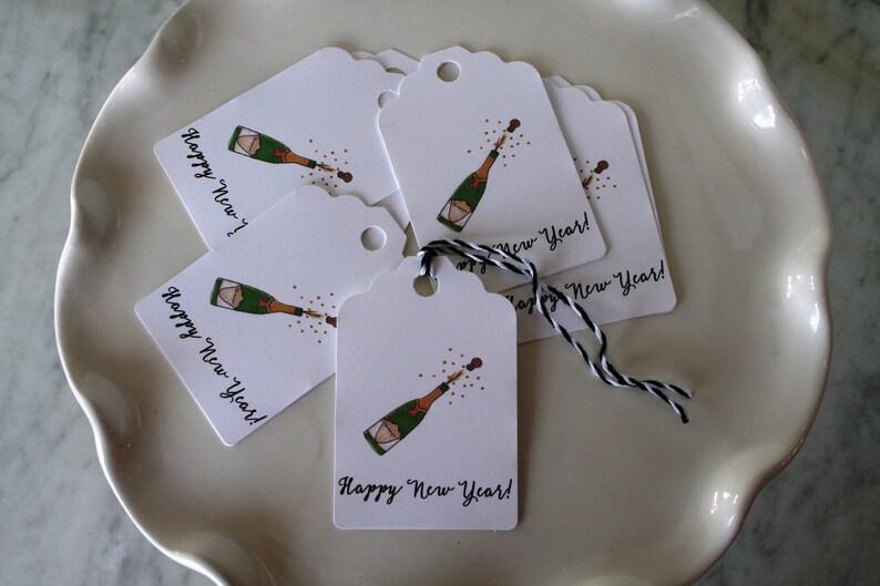 May include: A set of white gift tags with a black and white string. Each tag features a champagne bottle popping with the text "Happy New Year!" printed in black.