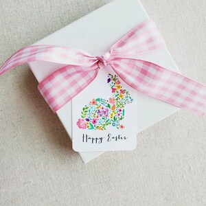 Puede incluir: Una caja de regalo blanca con una cinta de cuadros rosa atada en un lazo. Una etiqueta de regalo blanca con un dise&ntilde;o de conejo floral y el texto "Feliz Pascua" est&aacute; unida a la caja.