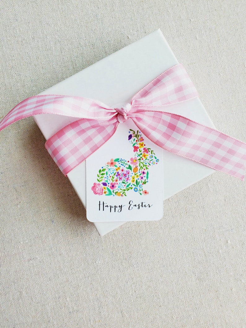 Puede incluir: Una caja de regalo blanca con una cinta de cuadros rosa atada en un lazo. Una etiqueta de regalo blanca con un dise&ntilde;o de conejo floral y el texto "Feliz Pascua" est&aacute; unida a la caja.
