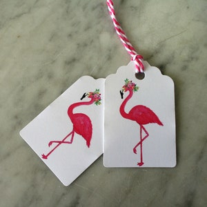 K&ouml;nnte beinhalten: Zwei Geschenkanh&auml;nger in Form von rosa Flamingos mit Blumenkr&auml;nzen. Die Anh&auml;nger sind wei&szlig; mit gewellten R&auml;ndern und einem Loch f&uuml;r eine Schnur.