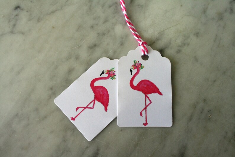 K&ouml;nnte beinhalten: Zwei Geschenkanh&auml;nger in Form von rosa Flamingos mit Blumenkr&auml;nzen. Die Anh&auml;nger sind wei&szlig; mit gewellten R&auml;ndern und einem Loch f&uuml;r eine Schnur.