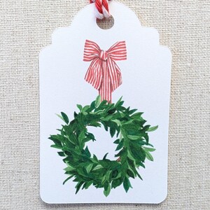 K&ouml;nnte beinhalten: Ein wei&szlig;es Geschenk-Etikett mit einer rot-wei&szlig; gestreiften Schleife und einer gr&uuml;nen Kranz-Illustration. Das Etikett ist mit rot-wei&szlig;em Garn versehen, ideal zum Verpacken von Weihnachtsgeschenken.