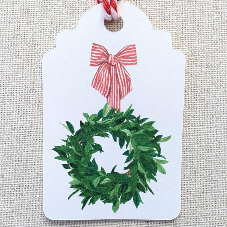 K&ouml;nnte beinhalten: Ein wei&szlig;es Geschenk-Etikett mit einer rot-wei&szlig; gestreiften Schleife und einer gr&uuml;nen Kranz-Illustration. Das Etikett ist mit rot-wei&szlig;em Garn versehen, ideal zum Verpacken von Weihnachtsgeschenken.