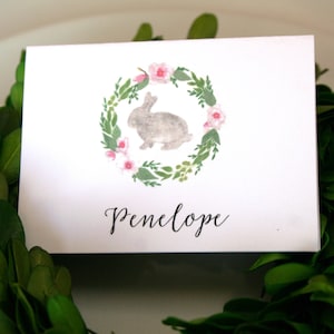 May include: A white card with a watercolor illustration of a bunny rabbit in a wreath of pink flowers. The text "Penelope" is written in black cursive below the illustration.