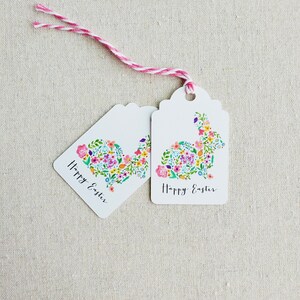 Puede incluir: Dos etiquetas de regalo blancas con un dise&ntilde;o de conejo floral y el texto "Feliz Pascua" en escritura cursiva negra. Las etiquetas est&aacute;n atadas juntas con un cordel rosa y blanco.