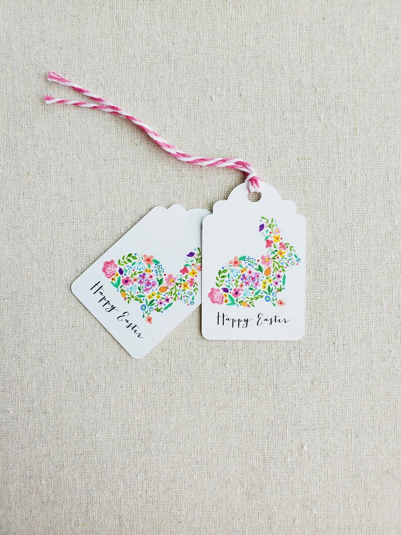 Puede incluir: Dos etiquetas de regalo blancas con un dise&ntilde;o de conejo floral y el texto "Feliz Pascua" en escritura cursiva negra. Las etiquetas est&aacute;n atadas juntas con un cordel rosa y blanco.