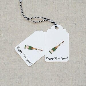 May include: Two white gift tags with a black and white twine string. Each tag features a hand-drawn illustration of a champagne bottle popping with the text "Happy New Year!"