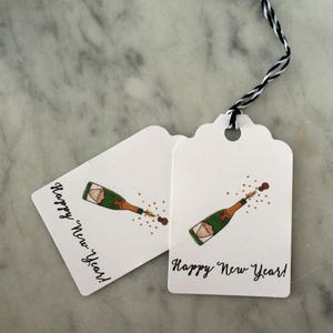 May include: Two white gift tags with black twine. Each tag features a hand-drawn illustration of a champagne bottle popping with the text "Happy New Year!"