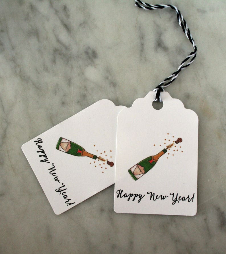May include: Two white gift tags with black twine. Each tag features a hand-drawn illustration of a champagne bottle popping with the text "Happy New Year!"