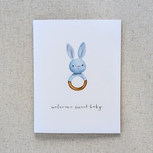 May include: A blue bunny rattle with a gold ring. The card reads "welcome sweet baby".