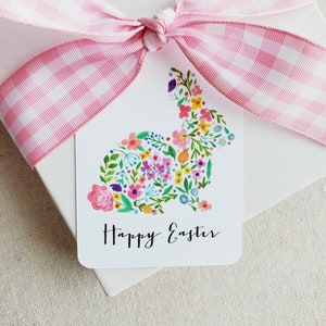 Puede incluir: Una etiqueta de regalo blanca con una cinta de gingham rosa y blanca atada en un lazo. La etiqueta presenta una silueta de conejo floral con el texto "Happy Easter" impreso debajo.