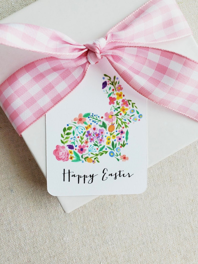 Puede incluir: Una etiqueta de regalo blanca con una cinta de gingham rosa y blanca atada en un lazo. La etiqueta presenta una silueta de conejo floral con el texto "Happy Easter" impreso debajo.