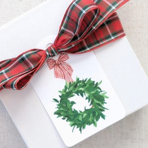 K&ouml;nnte beinhalten: Eine wei&szlig;e Geschenkbox, gebunden mit einem rot-gr&uuml;nen Karoband. Ein Geschenkanh&auml;nger mit einem Aquarell-Kranz-Design und einer rot-wei&szlig; gestreiften Schleife ist am Band befestigt.