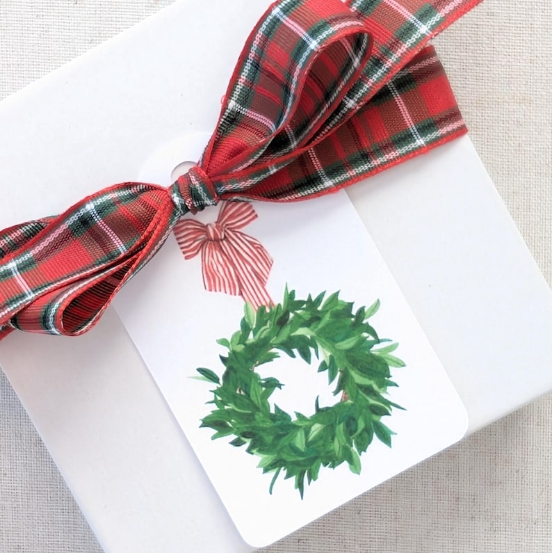 K&ouml;nnte beinhalten: Eine wei&szlig;e Geschenkbox, gebunden mit einem rot-gr&uuml;nen Karoband. Ein Geschenkanh&auml;nger mit einem Aquarell-Kranz-Design und einer rot-wei&szlig; gestreiften Schleife ist am Band befestigt.