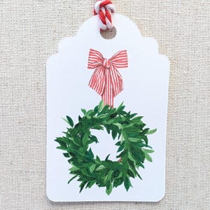 K&ouml;nnte beinhalten: Ein wei&szlig;es Geschenk-Etikett mit einer rot-wei&szlig; gestreiften Schleife und einer gr&uuml;nen Kranz-Illustration. Das Etikett ist mit rot-wei&szlig;em Garn versehen. Ideal f&uuml;r Weihnachts- oder Urlaubsgeschenke.