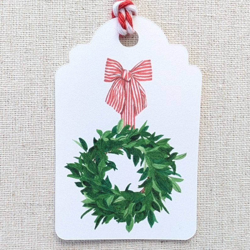 K&ouml;nnte beinhalten: Ein wei&szlig;es Geschenk-Etikett mit einer rot-wei&szlig; gestreiften Schleife und einer gr&uuml;nen Kranz-Illustration. Das Etikett ist mit rot-wei&szlig;em Garn versehen. Ideal f&uuml;r Weihnachts- oder Urlaubsgeschenke.