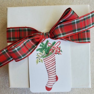 May include: A white gift box with a red and green plaid ribbon tied in a bow. A white gift tag with a red and white striped stocking with green holly and red berries is attached to the ribbon.